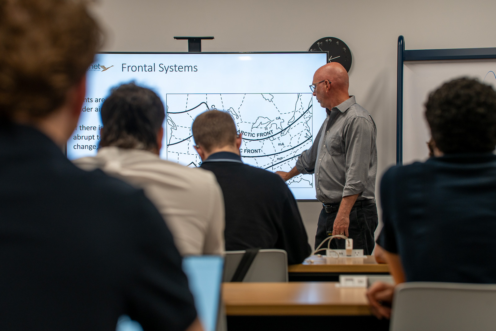 A presenter stands at the front of a classroom, pointing to a weather map displayed on a large screen titled “Frontal Systems.” Several attendees are seated at desks in the foreground, viewed from behind, while the presentation focuses on atmospheric fronts and weather patterns. The room has a training or workshop setting with a whiteboard and wall clock visible in the background.